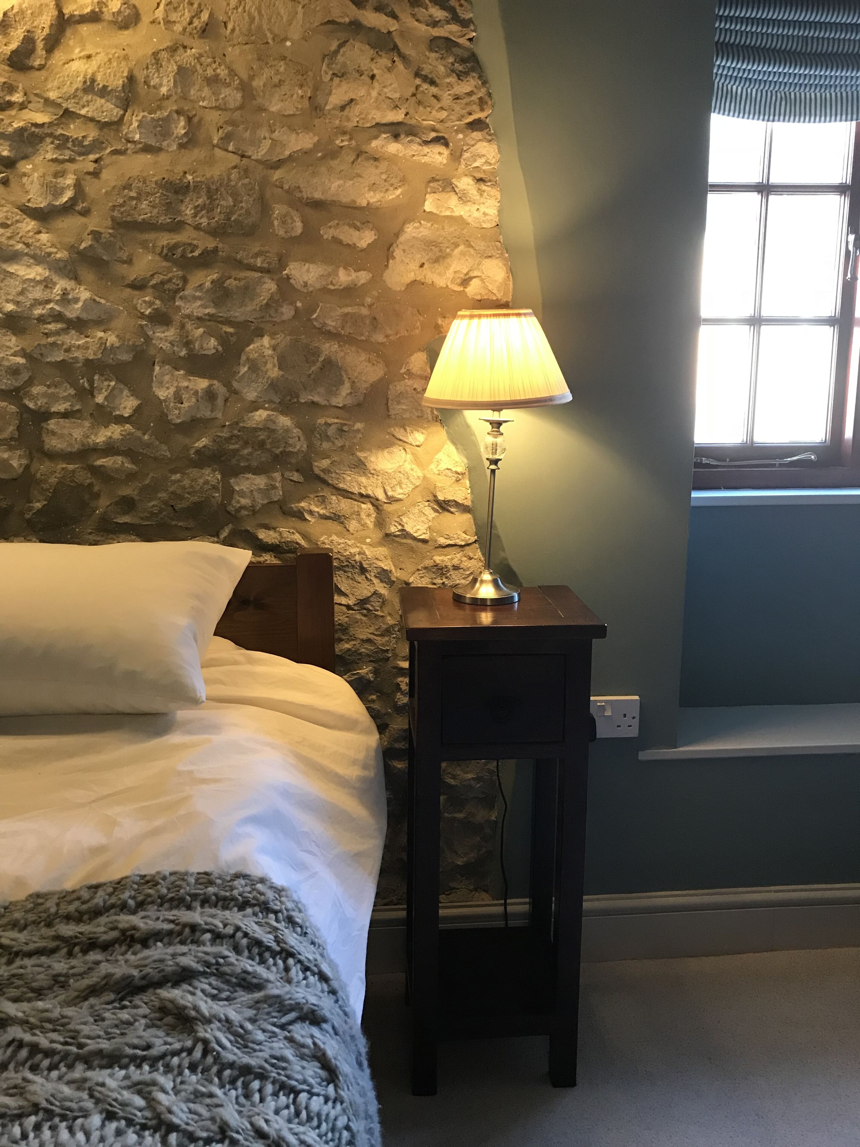 Een slaapkamer met een stenen muur, een bed met wit beddengoed, een lamp op een nachtkastje en een raam met jaloezieën.