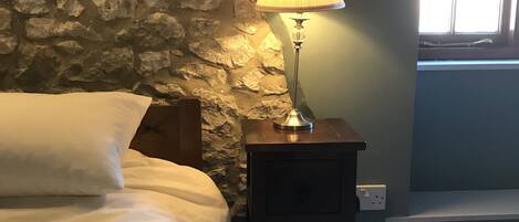 Een slaapkamer met een stenen muur, een bed met wit beddengoed, een lamp op een nachtkastje en een raam met jaloezieën.