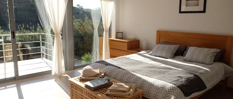 Ein Schlafzimmer mit einem Bett, einem Nachttisch, Vorhängen an einem Fenster und Blick auf Grün außerhalb.
