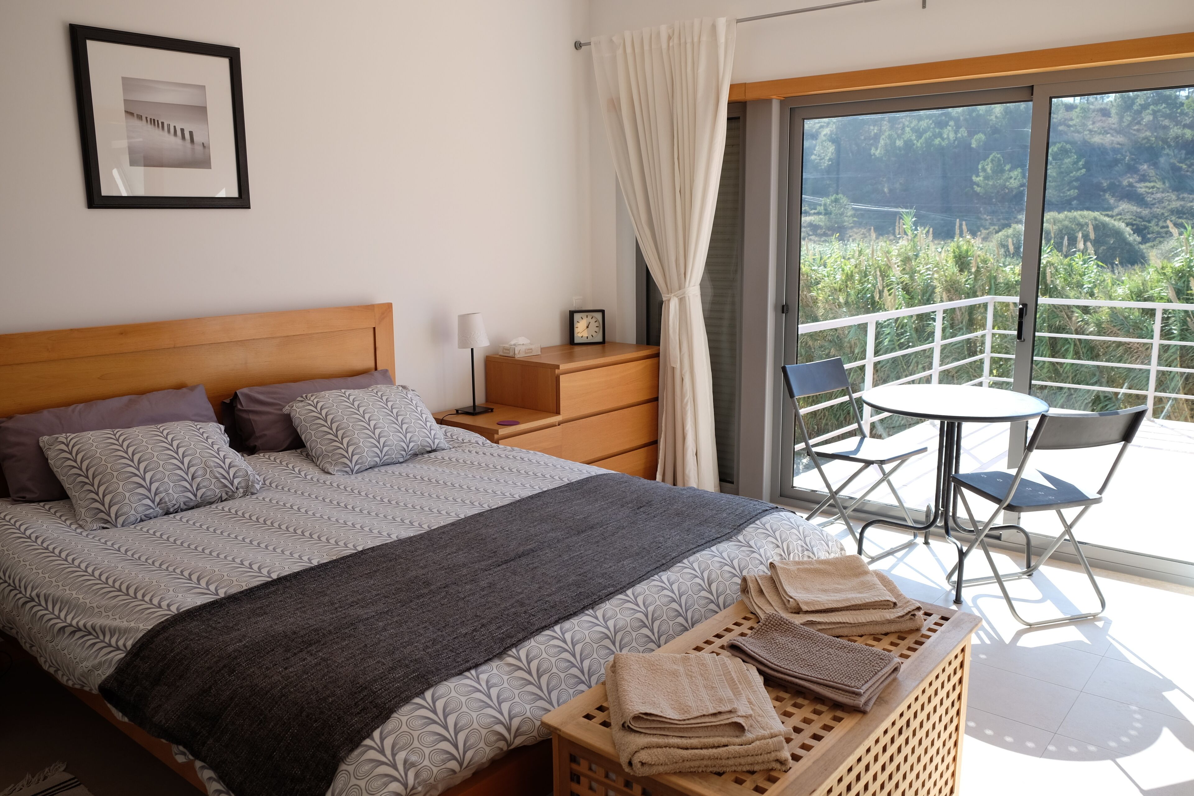 A bedroom with a bed, a nightstand, a bedside table, a window with curtains, and a balcony with a table and chairs.