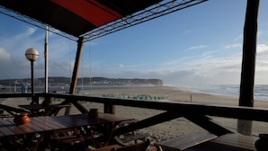 An outdoor seating area with wooden tables and chairs, overlooking a beach with a clear view of the ocean and distant hills.
