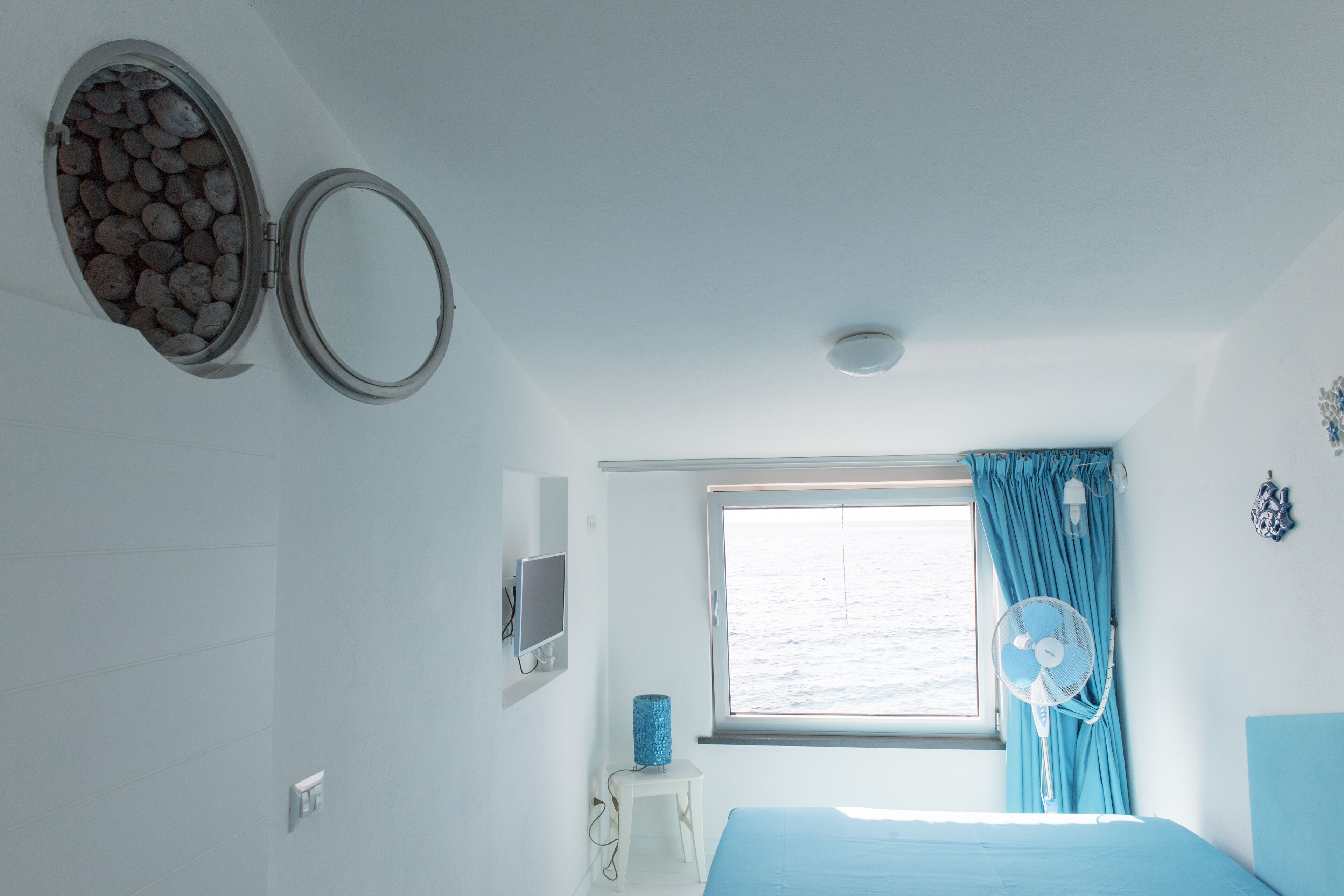 Ein Schlafzimmer mit Bett, Fenster mit Vorhängen, einem Ventilator und einem an der Wand befestigten Spiegel.