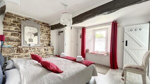 Un dormitorio con pared de piedra, cama con almohadas rojas, una silla blanca y una ventana con cortinas rojas.