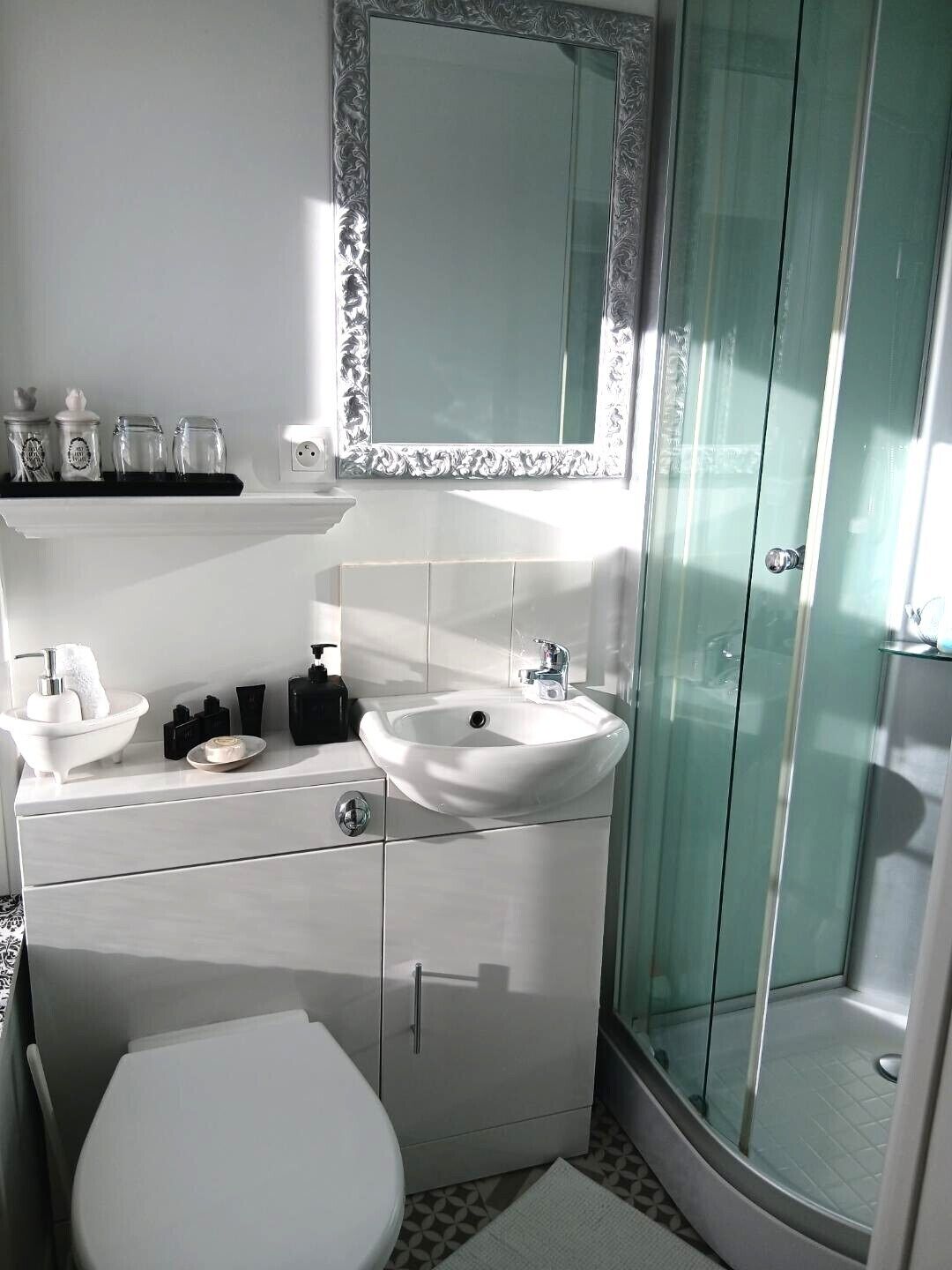Une salle de bain moderne avec un lavabo blanc, un miroir et des toilettes. On y trouve une douche en verre et une étagère avec des articles de toilette.