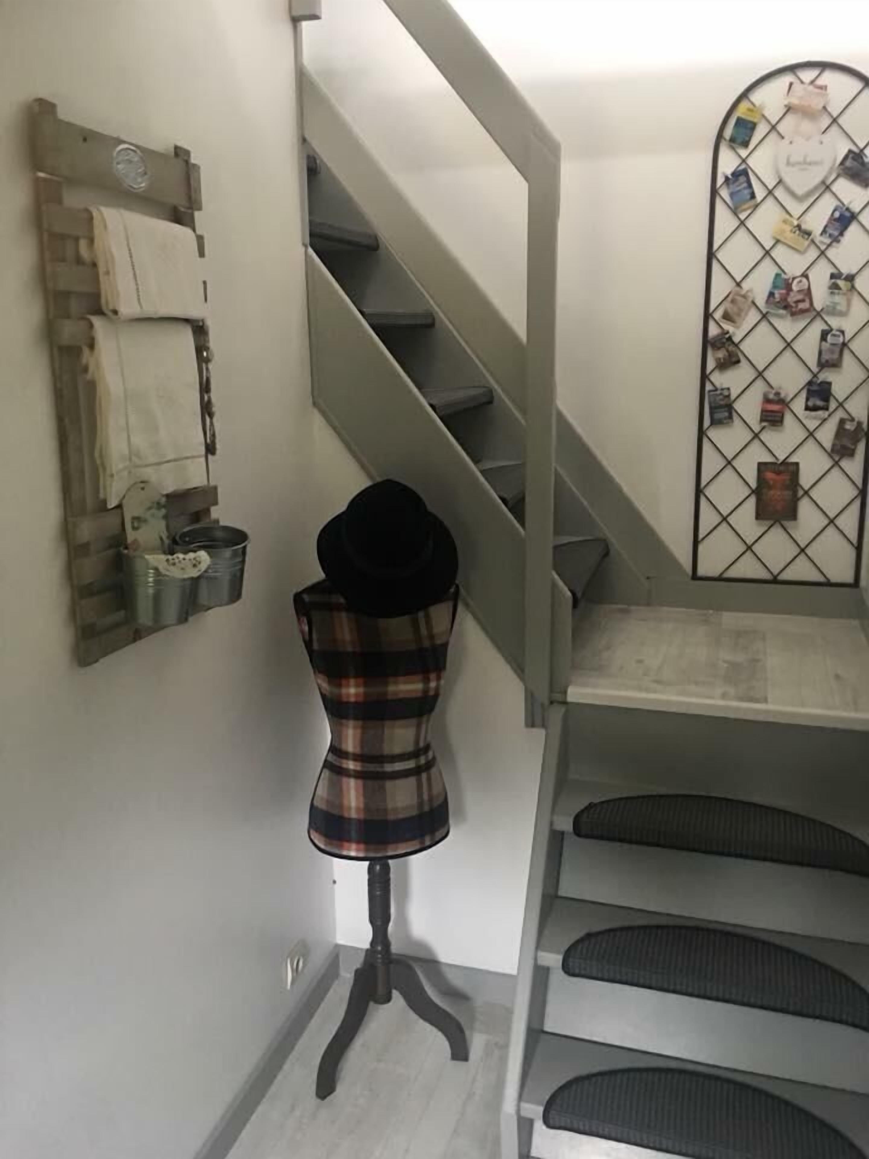 Una escalera con barandilla de metal, un maniquí vestido con un traje a cuadros, y un estante en la pared con diversos objetos.