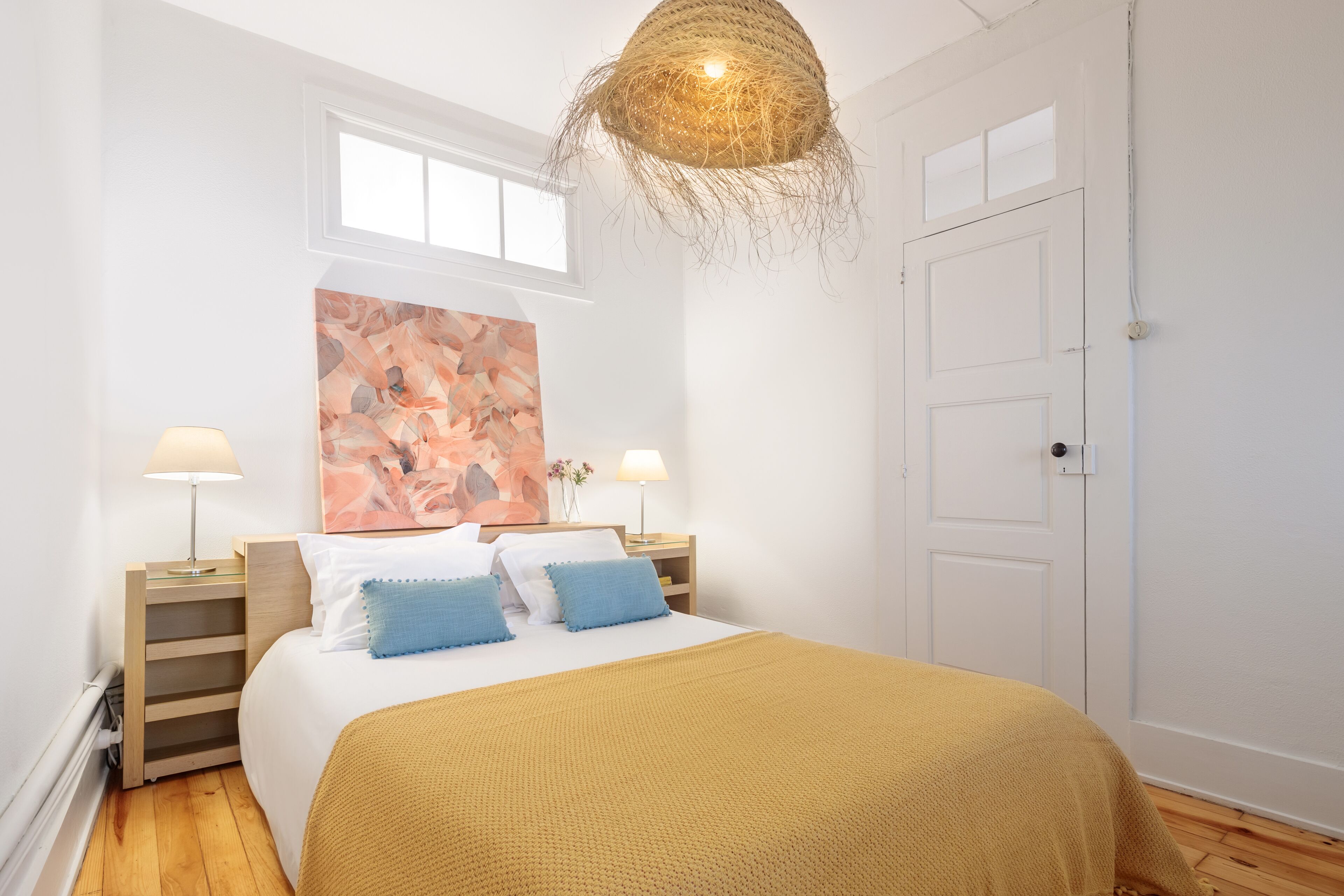 A bedroom with a bed, two bedside tables with lamps, a large abstract painting on the wall, and a door.