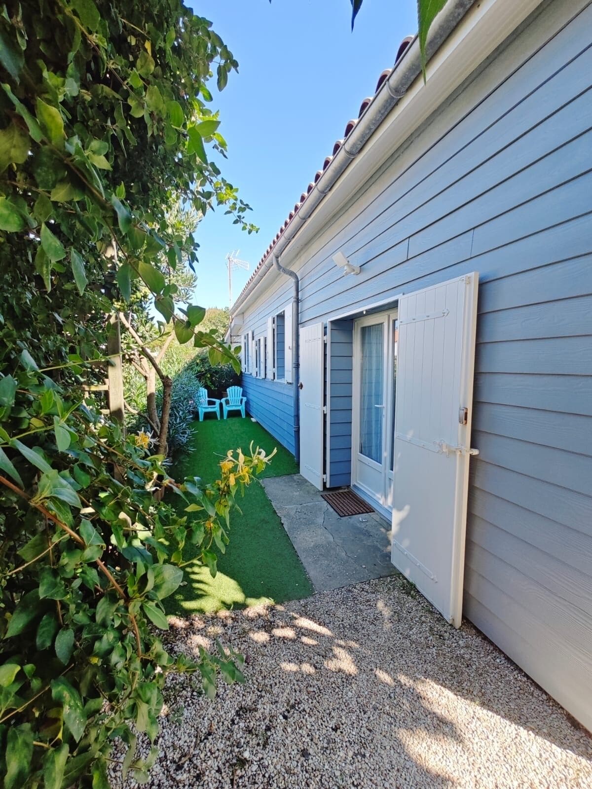 Ein schmaler Weg führt zu einem Gebäude mit blauer Fassade und einem kleinen Gartenbereich mit Kunstgrashalm und Kiesweg.