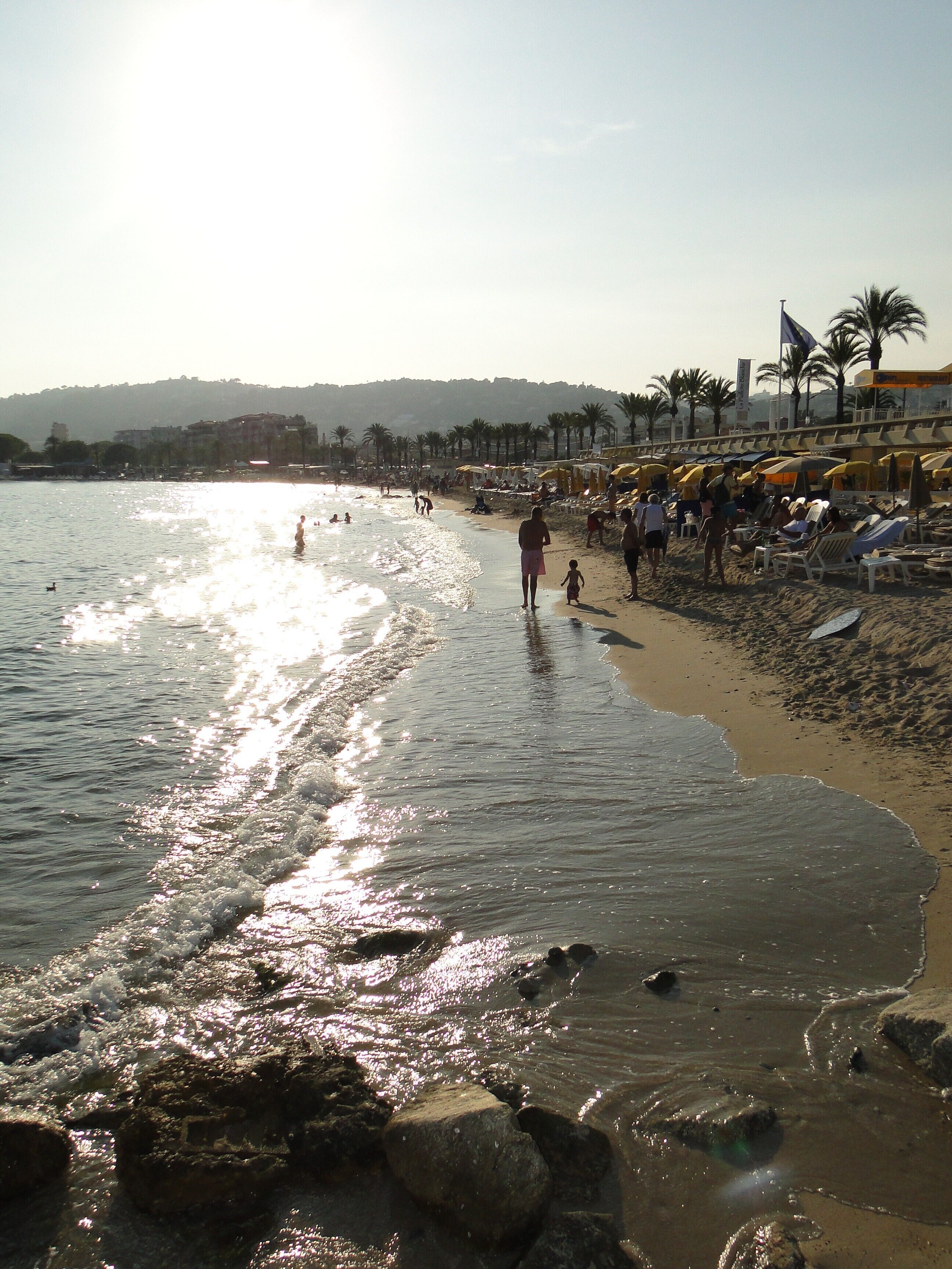 A beach with people walking along the shore, umbrellas, and palm trees in the background.