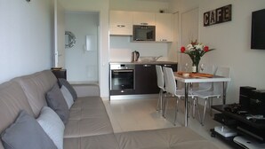 A modern living room with a sofa, a dining table with chairs, a kitchen area with appliances, and a flat-screen TV mounted on the wall. - 2 bedroom, Overlooking Parc Exflora + Sea views. Pool+Beach 200m + Parking + A/C (Antibes)