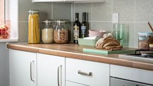 A kitchen with white cabinets, a wooden countertop, and various food items including jars, bottles, and bread. - Deco Spacious Townhouse (Coventry)