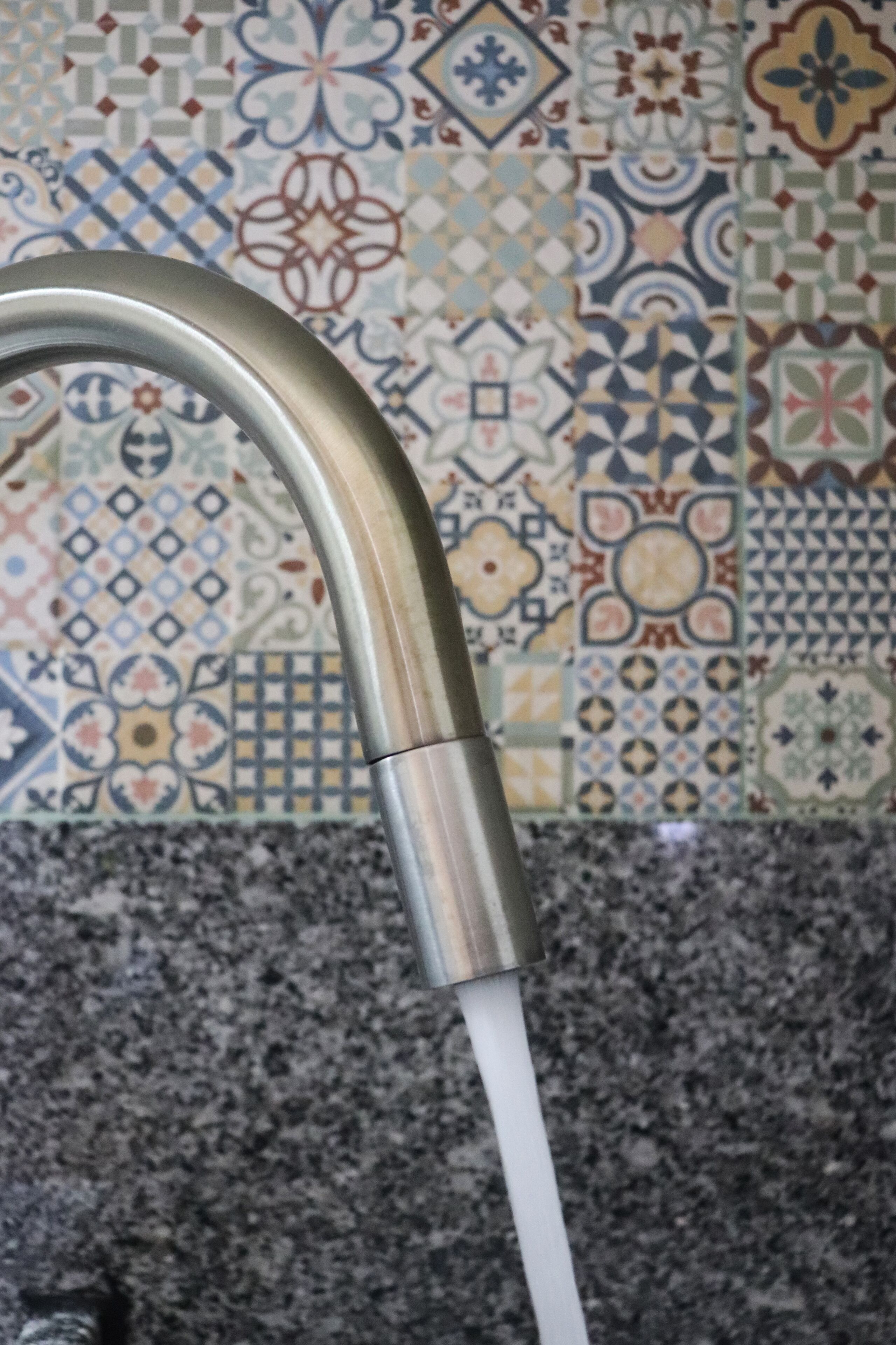 A faucet with water running, set against a tiled wall with a variety of patterns.