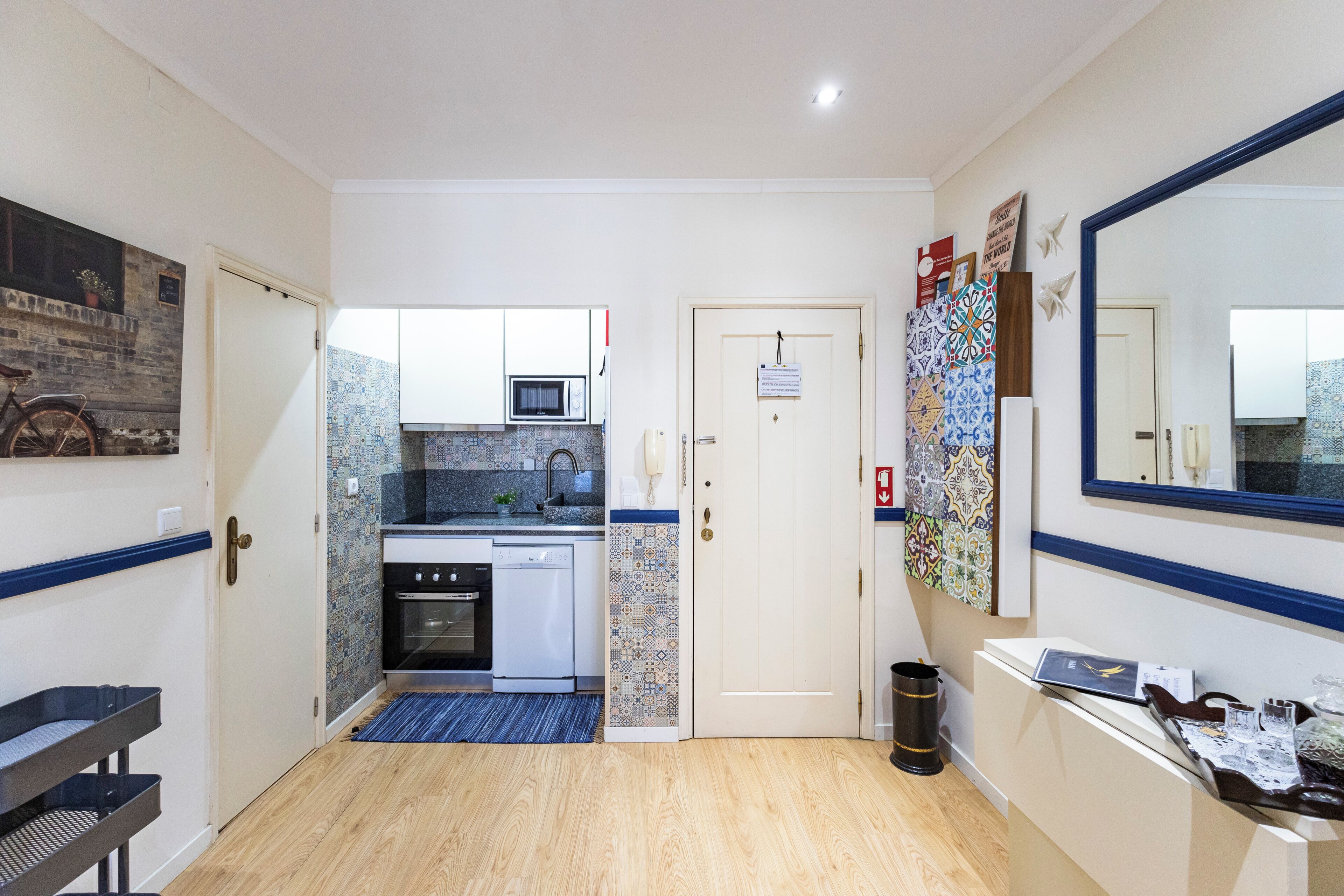 A hallway leading to a kitchen with a microwave, oven, and dishwasher. There is a door with a sign, a trash can, and a bench.