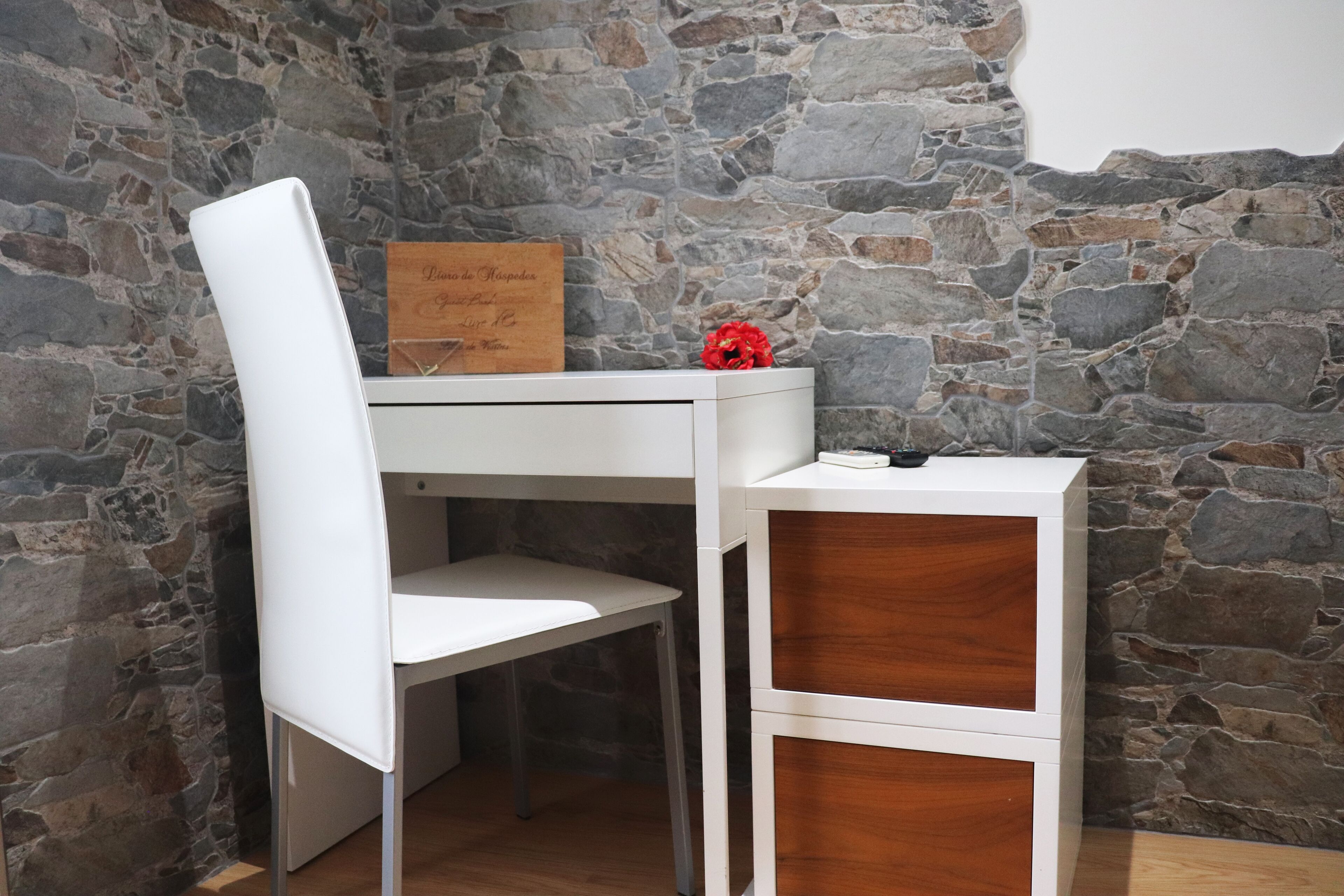 Ein weißer Schreibtisch mit Stuhl, ein kleines Holzschränkchen und eine Steinwand.