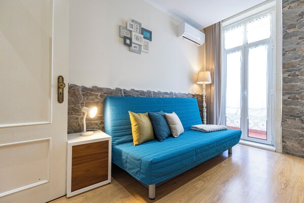 A room with a blue sofa, a bedside lamp, a stone accent wall, a white door, and a window with curtains.