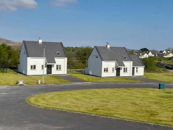 Two charming homes with distinct entrances and porches sit amidst lush greenery and rolling hills.