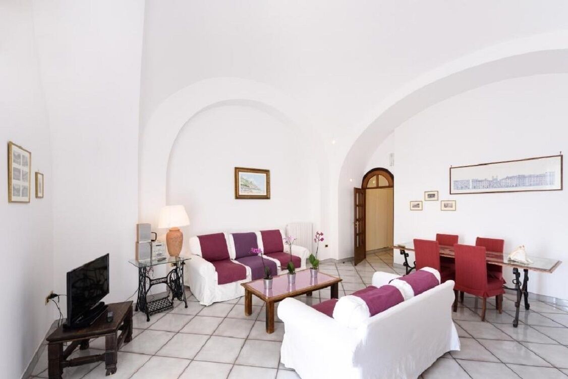 A spacious living room with white sofas, a coffee table, and a dining area with red chairs.