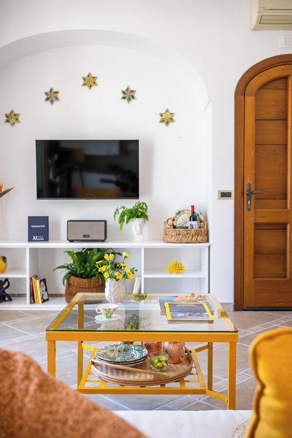A living room with a glass-top coffee table, a television mounted on the wall, a wooden door, and a white wall with star decorations. - Cozy house with sea view, terrace and private parking (Positano)