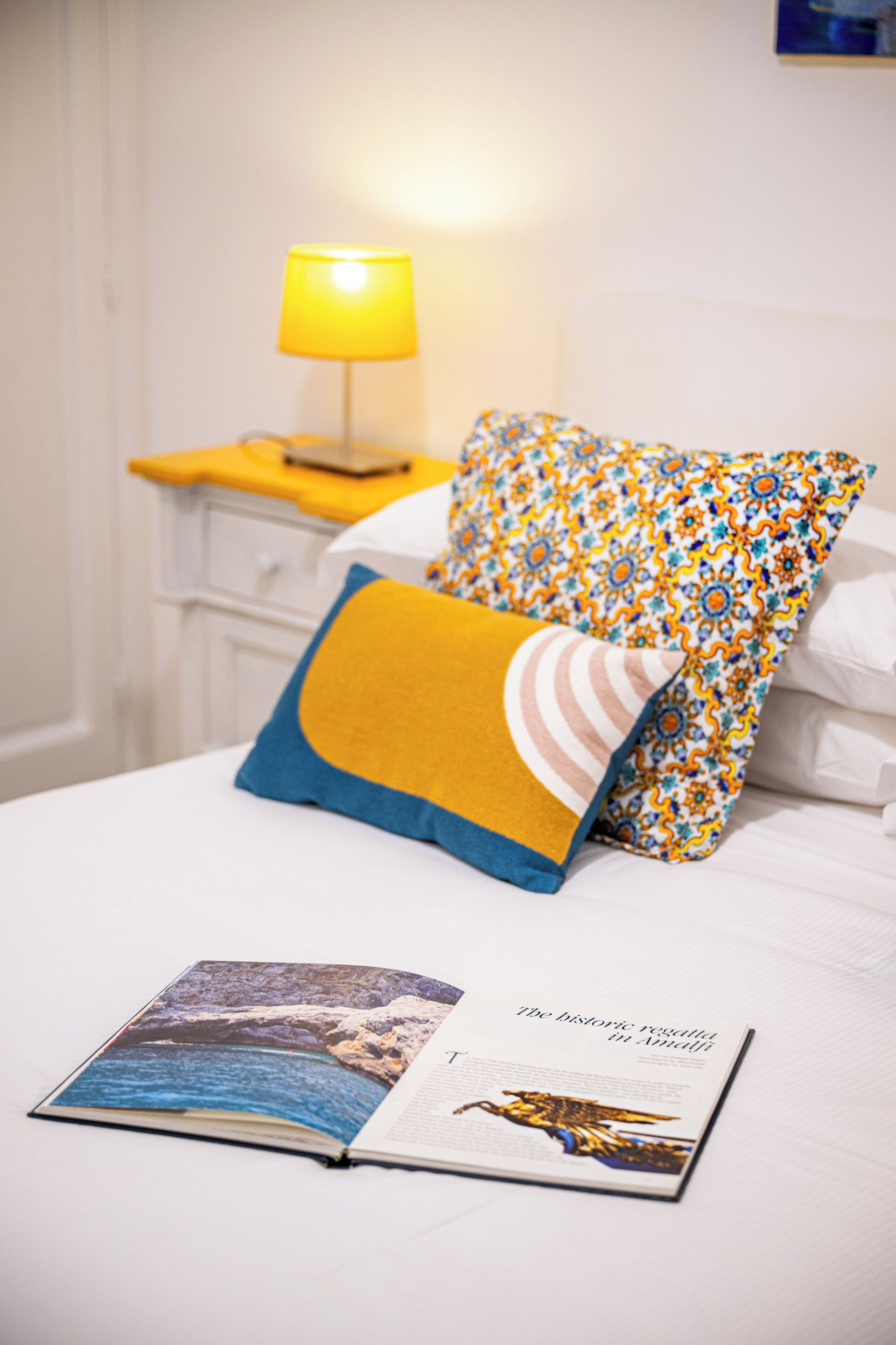 A neatly made bed with a magazine on it, a yellow lamp on a bedside table, and patterned pillows.