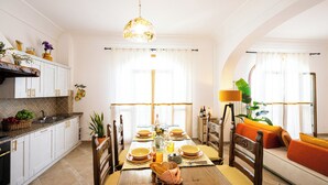 Un coin repas avec une table en bois, dressée pour quatre personnes, entourée de chaises. On y trouve également une cuisine avec des placards blancs et un évier, situés à gauche.