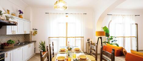 A dining area with a wooden table set for four, surrounded by chairs. There is a kitchen area with white cabinets and a sink to the left.