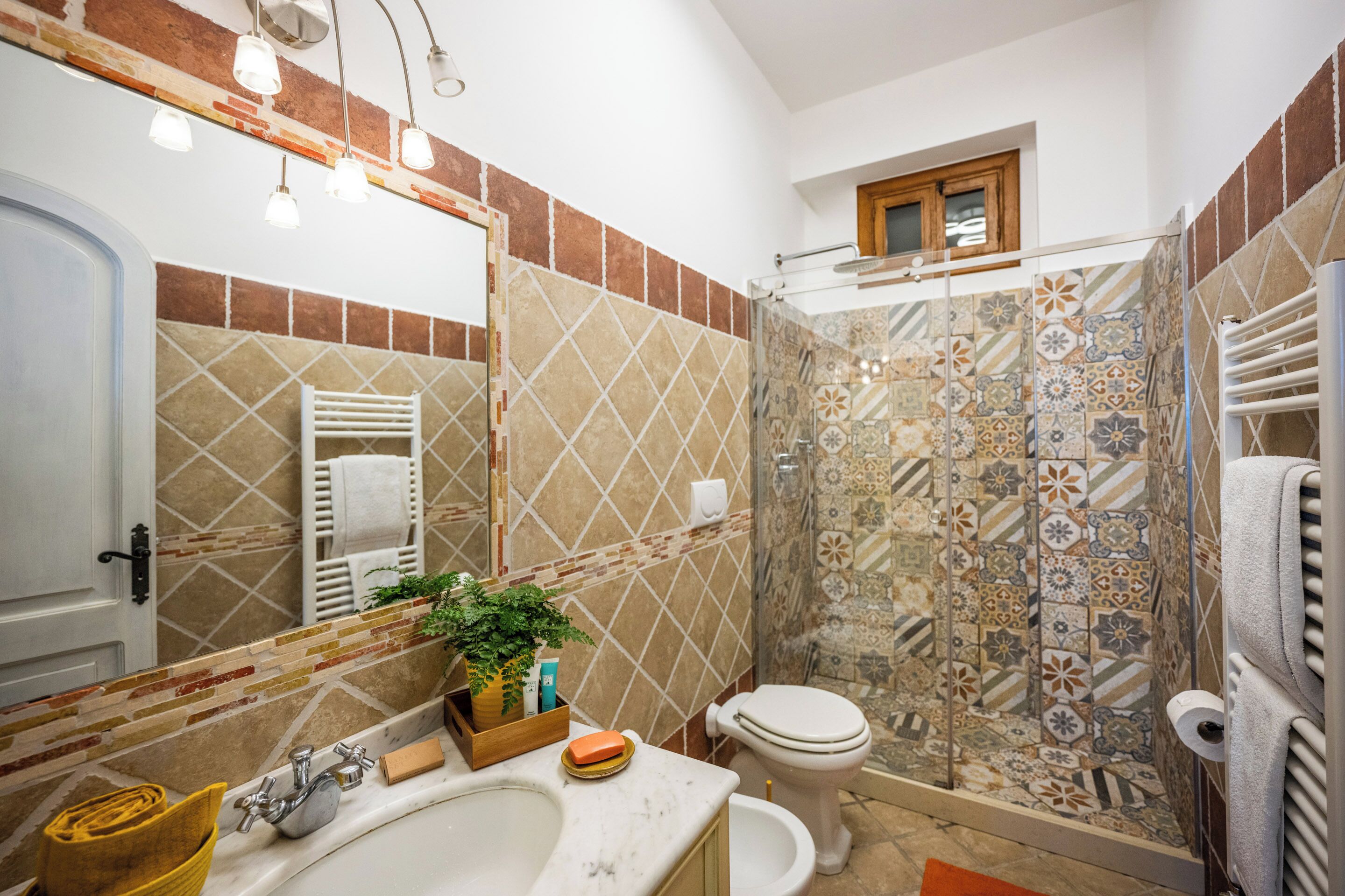 Un baño con lavamanos de mármol, inodoro, ducha con azulejos estampados y espejo con iluminación.
