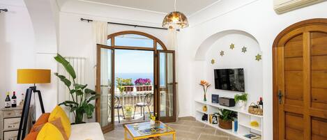 A living room with a sofa, a coffee table, a TV, and a balcony with plants and a view.