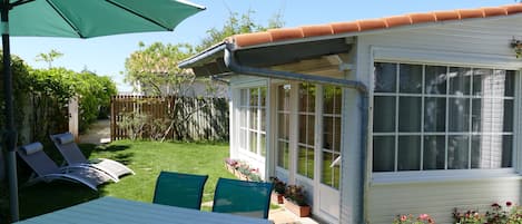 Un patio con una mesa y sillas, un espacio cubierto con una sombrilla, y una casa pequeña con una ventana.
