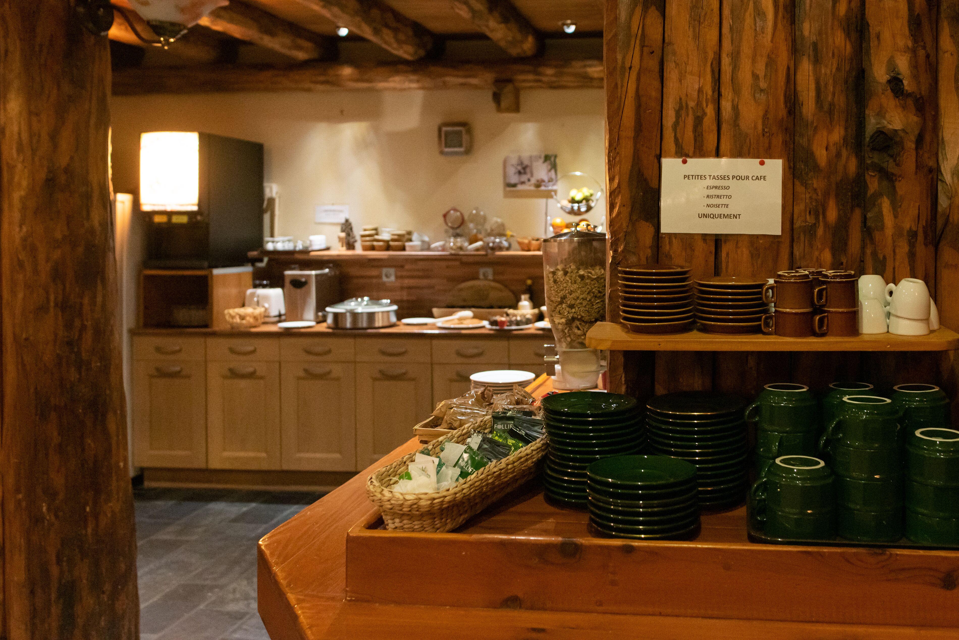 A breakfast buffet area with green plates, mugs, and a sign listing available coffee options.