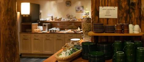 A breakfast buffet area with green plates, mugs, and a sign listing available coffee options.