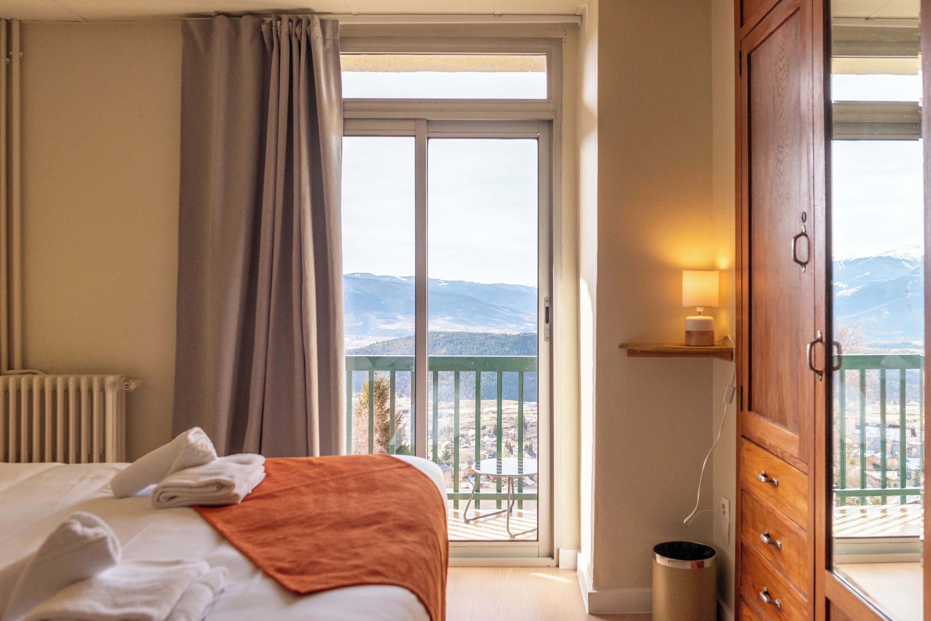 A hotel room with a bed, a balcony with a view of mountains, a wooden cabinet, and a lamp.