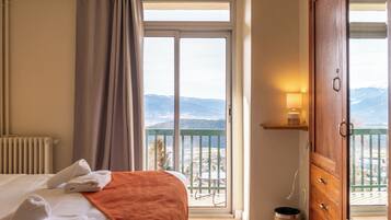 A hotel room with a bed, a balcony with a view of mountains, a wooden cabinet, and a lamp.
