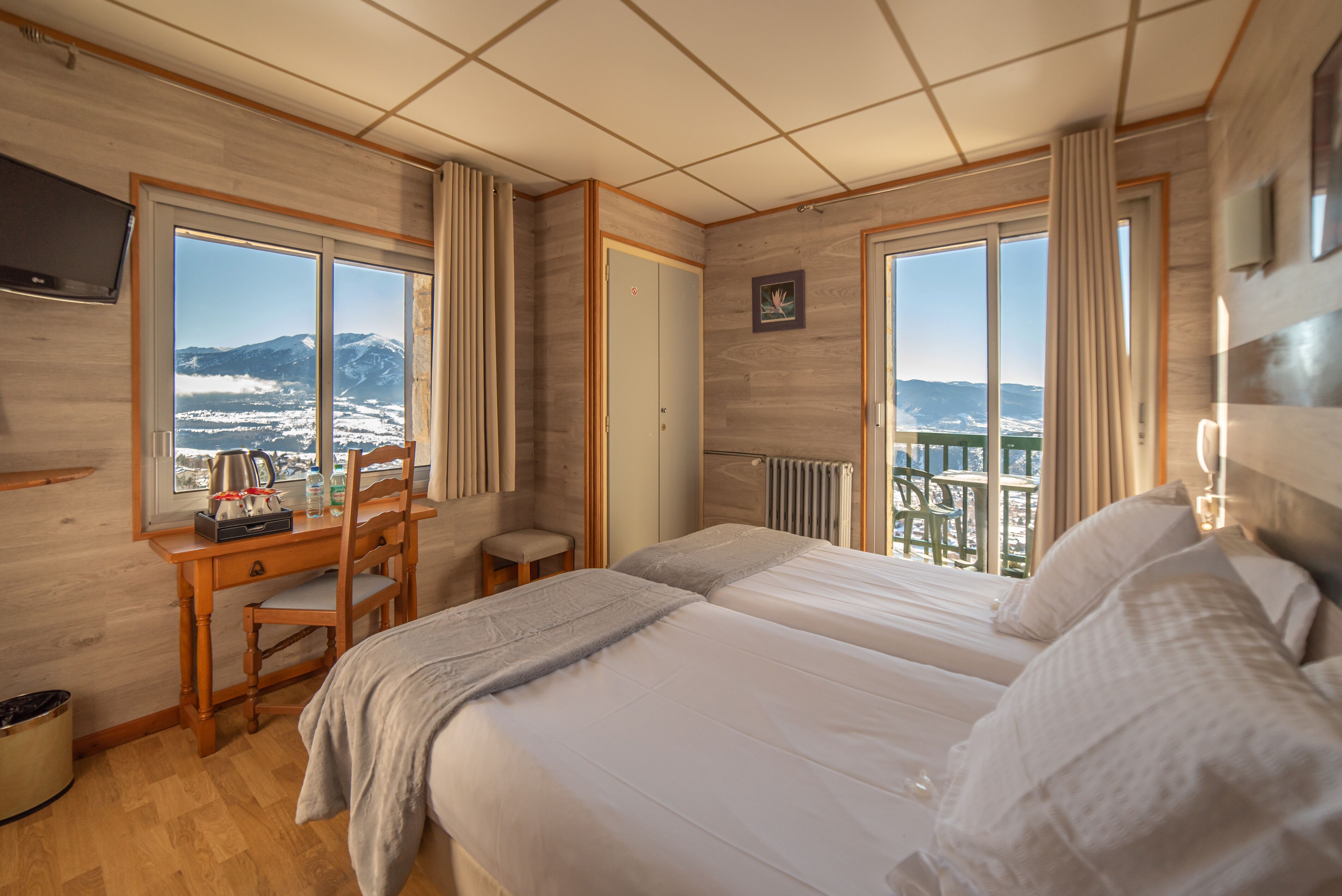 A hotel room with a wooden interior, a desk with a chair, a bed, and large windows offering a mountain view.