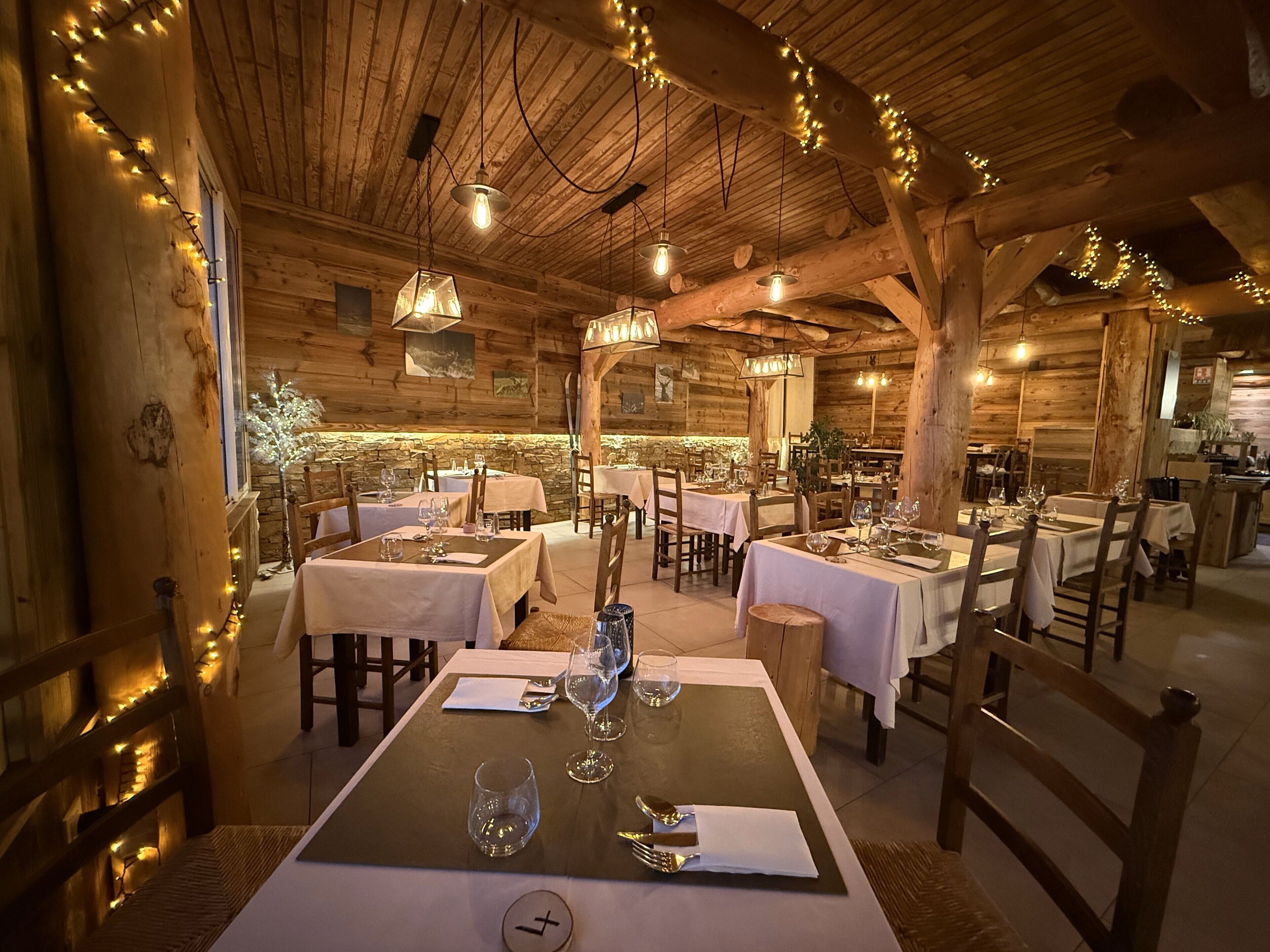 A cozy, rustic wooden interior with tables set for dining, string lights, and a stone wall.