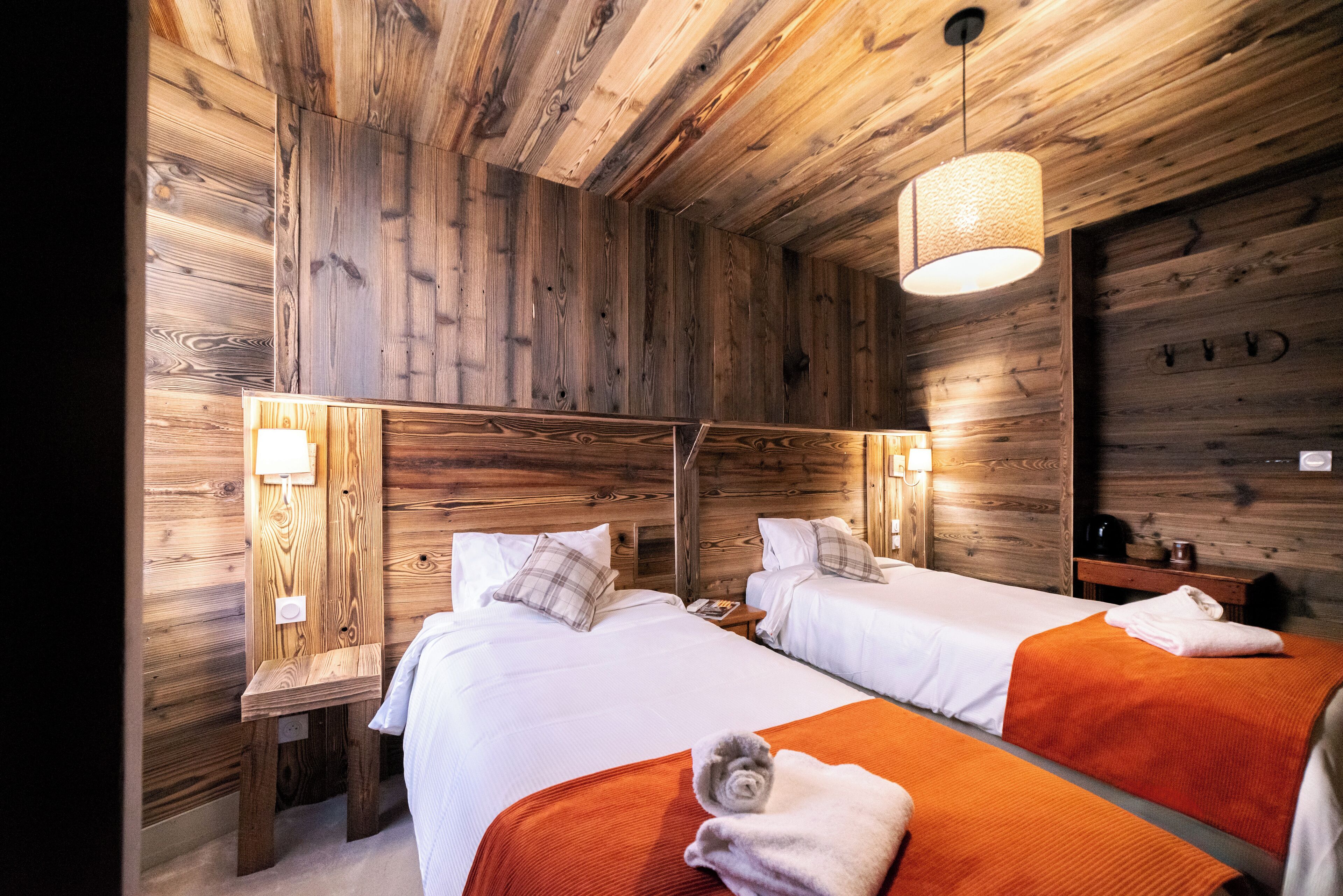 Una habitación tipo cabaña de madera con dos camas, cada una con cubrecama blanco y mantas naranjas. Tiene cabeceras de madera, una pequeña mesita de noche con lámpara y una lámpara colgante.