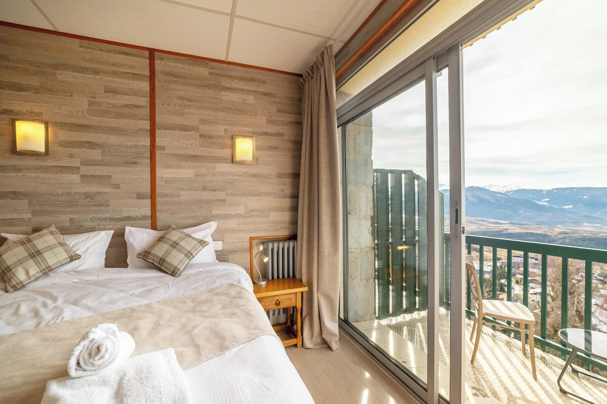 A hotel room with a large bed, a wooden headboard, a desk with a lamp, and a balcony with a view of mountains.