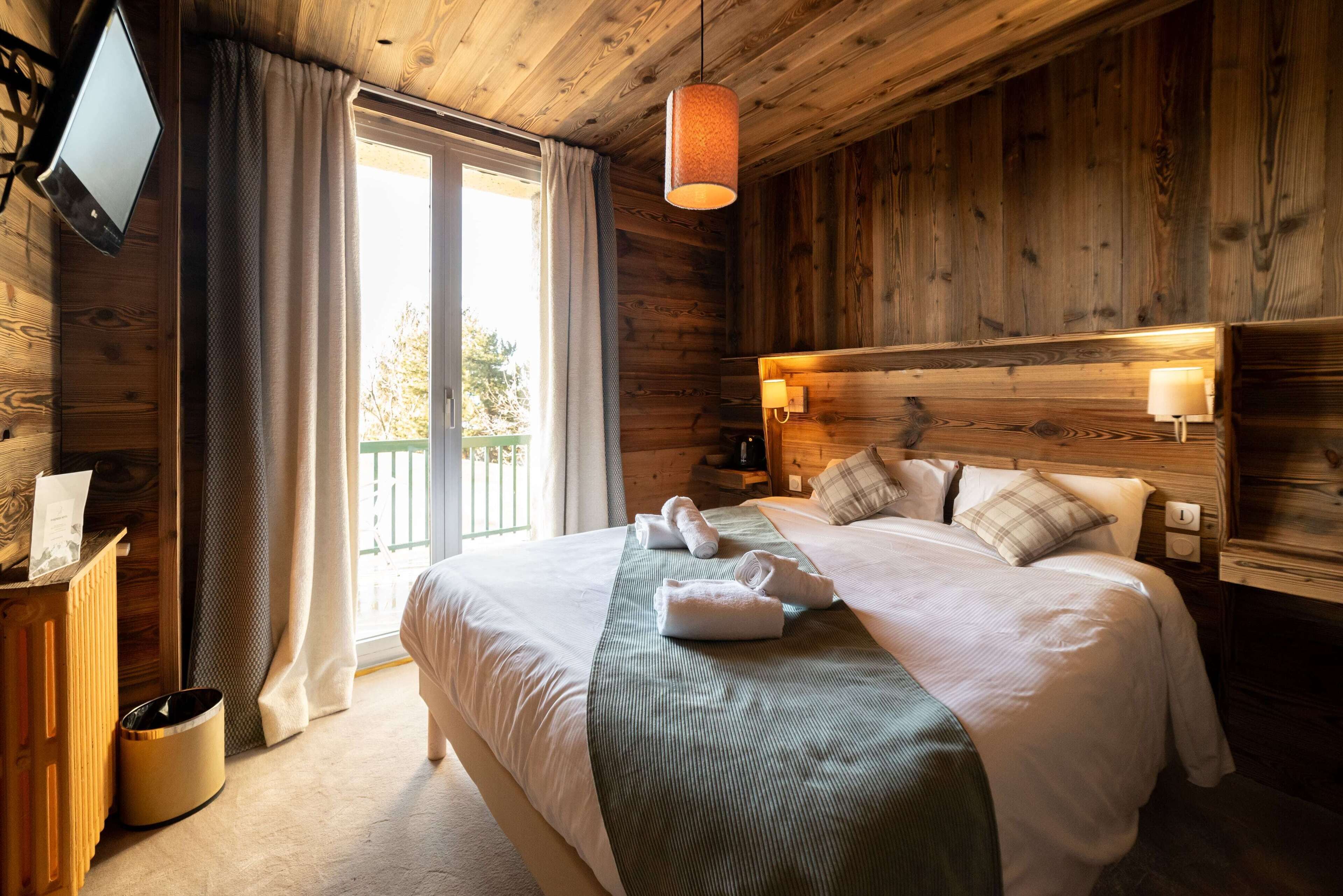 A wooden cabin-style bedroom with a large bed, a TV mounted on the wall, and a balcony visible through the window.