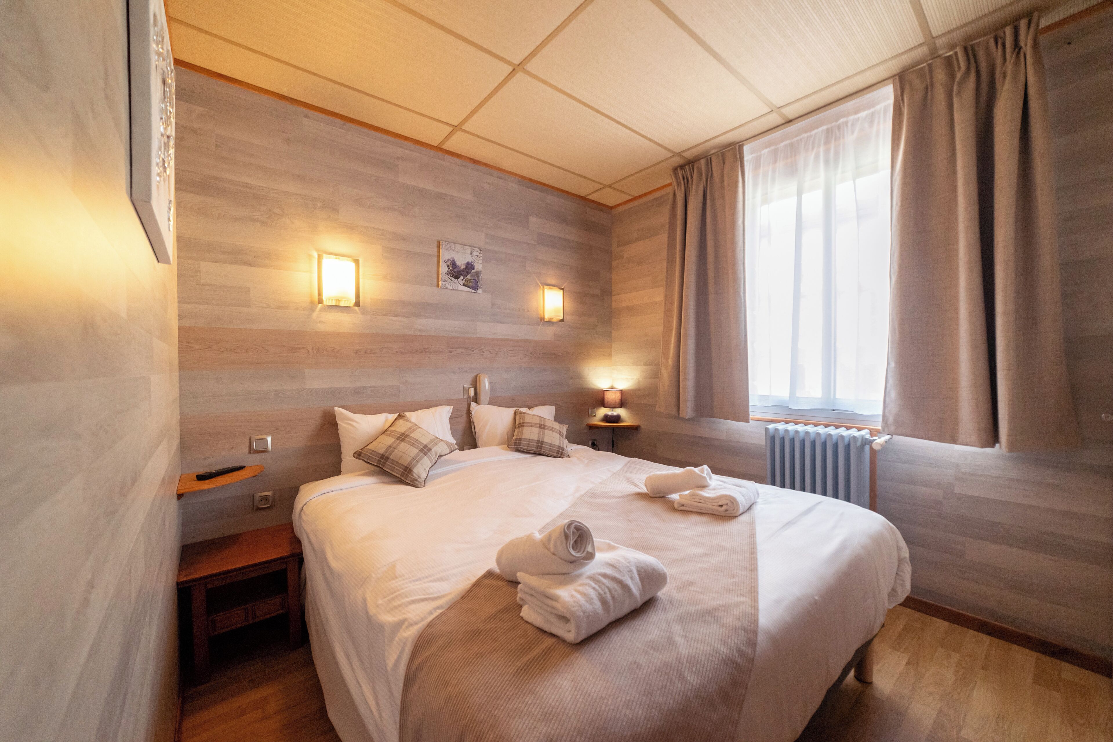 Una cama bien hecha con toallas, una mesita de noche y una lámpara de pared, junto a una ventana con cortinas.