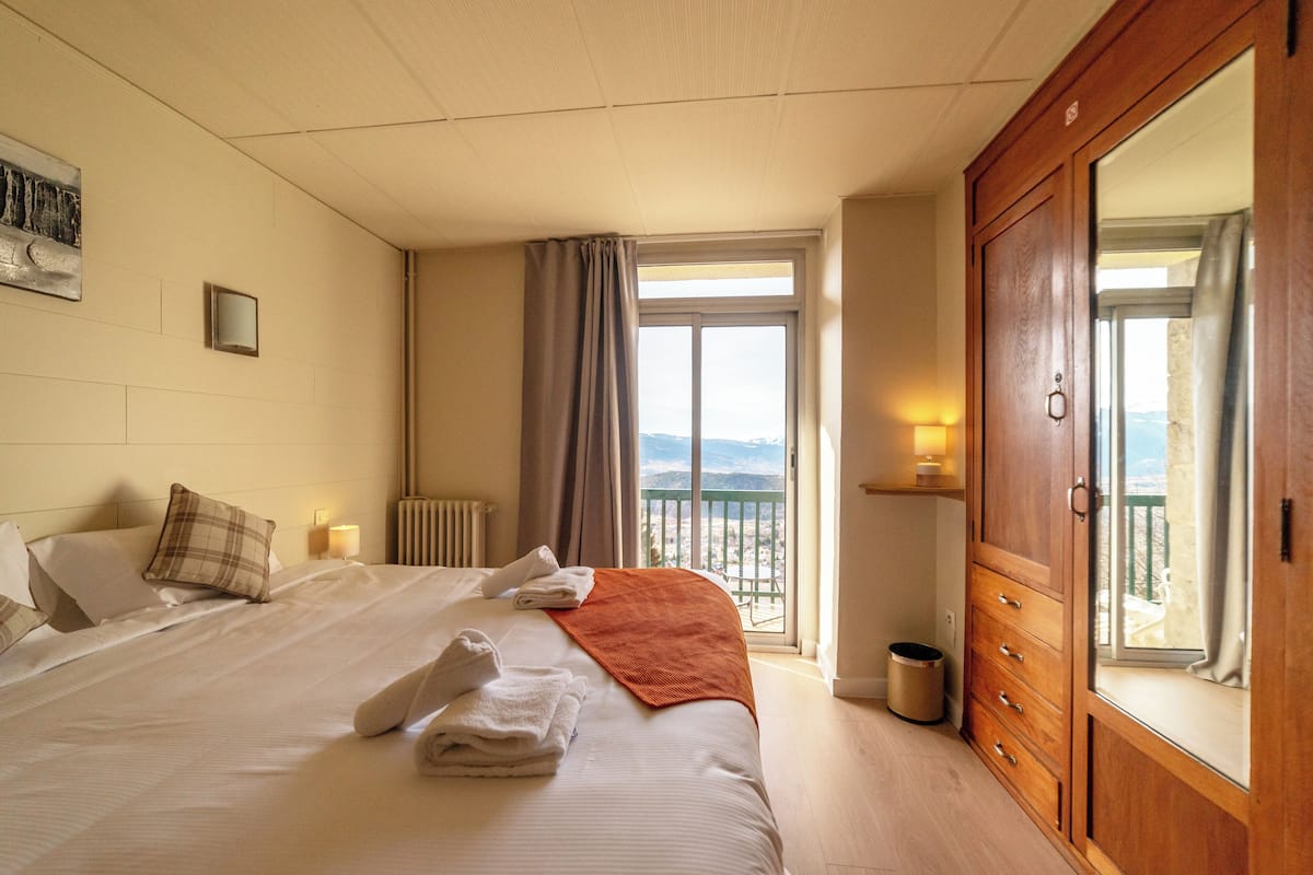 A hotel room with a large bed, a wooden wardrobe, a balcony with a view, and a window with curtains.