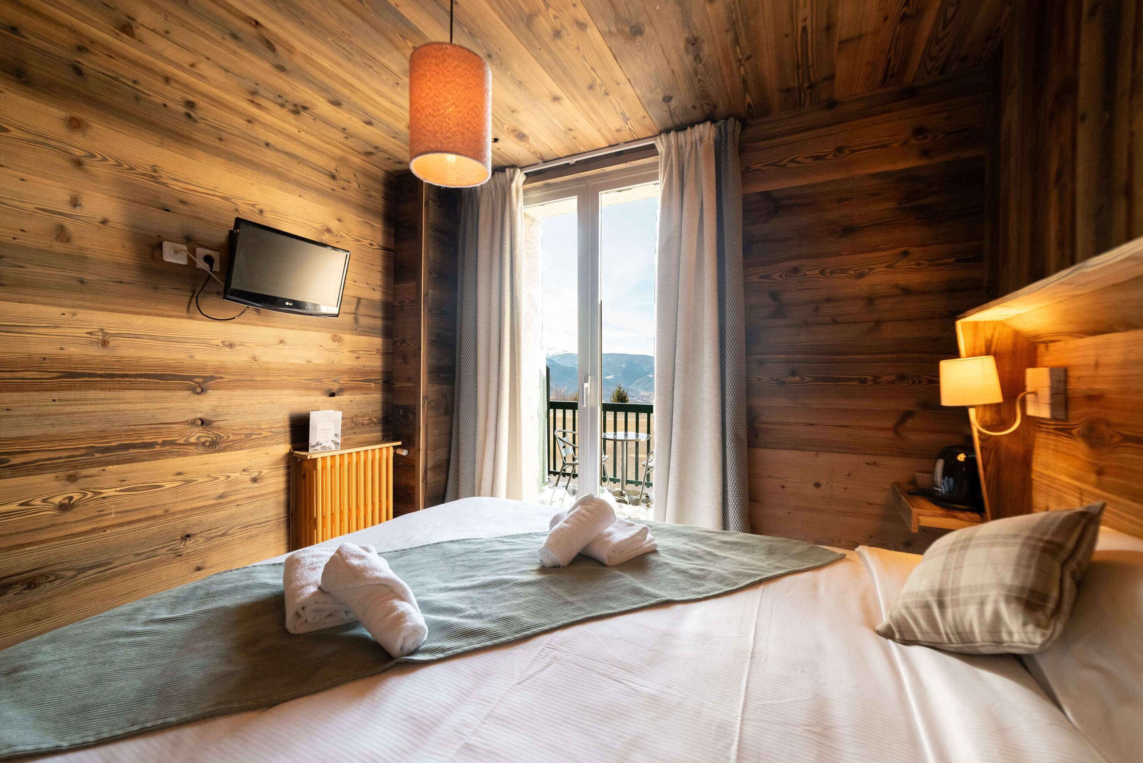 A wooden cabin-style room with a bed, a TV mounted on the wall, and a view of the outdoors through a window.