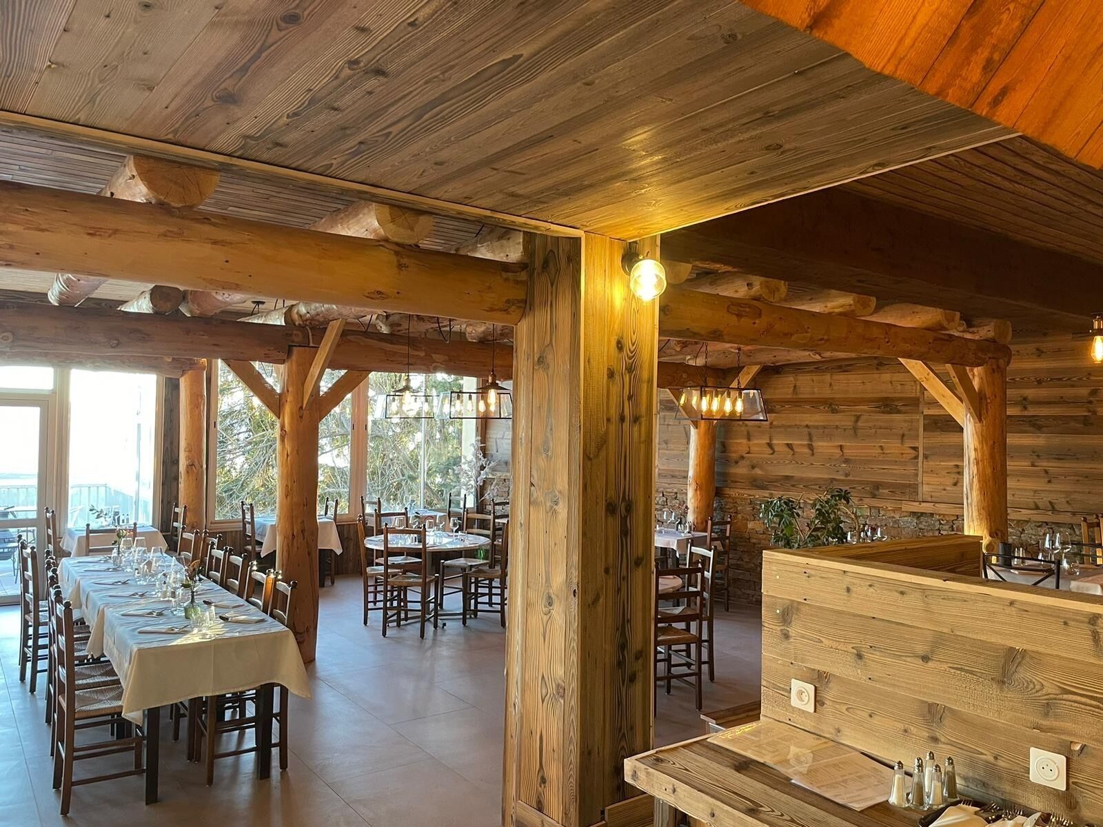 A rustic wooden interior with multiple dining tables and chairs, large windows, and a high ceiling with exposed beams.