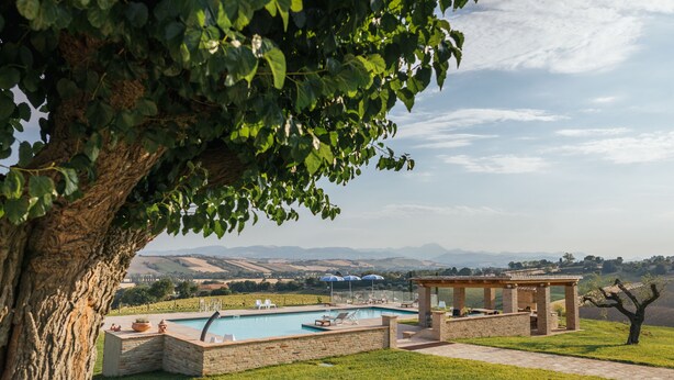 A pool area with lounge chairs and a pergola, surrounded by a grassy landscape and a view of distant hills.