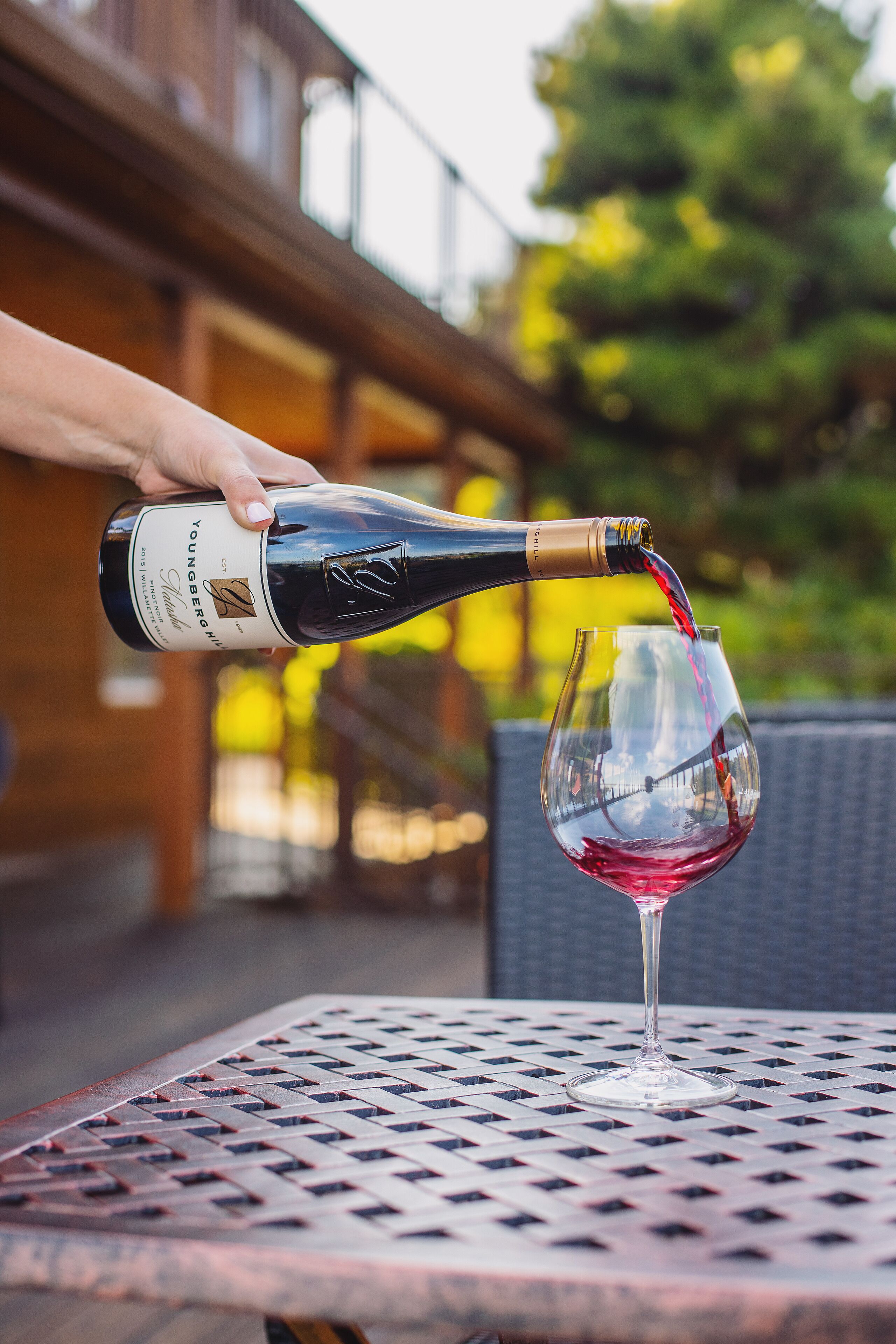 A hand pouring wine from a bottle into a glass on a metal table outdoors.