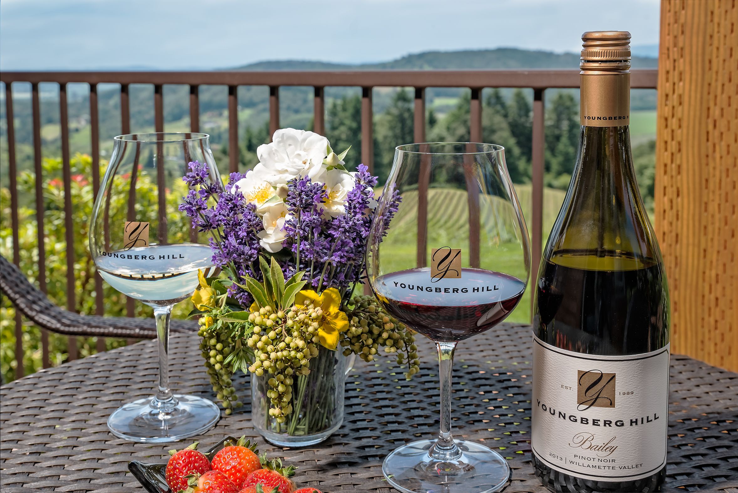 Une bouteille de Youngberg Hill Bailey Pinot Noir est posée sur une table, accompagnée de deux verres à vin, dont l’un est rempli de vin blanc et l’autre de vin rouge, ainsi qu’un vase de fleurs.