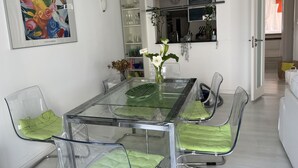 Ein Essbereich mit einem Glastisch und Stühlen, einem gerahmten Gemälde sowie einer Küchenzeile mit einer Uhr und einem Fenster.
