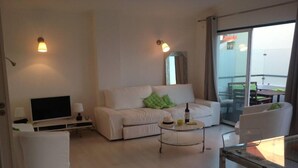 Ein modernes Wohnzimmer mit einer Couch, einem Fernseher, einem Glastisch und einem Balkon mit Gartenmöbeln.