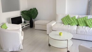 Ein modernes Wohnzimmer mit einem weißen Sofa, einem weißen Sessel, einem runden Glastisch und einer großen Zimmerpflanze.