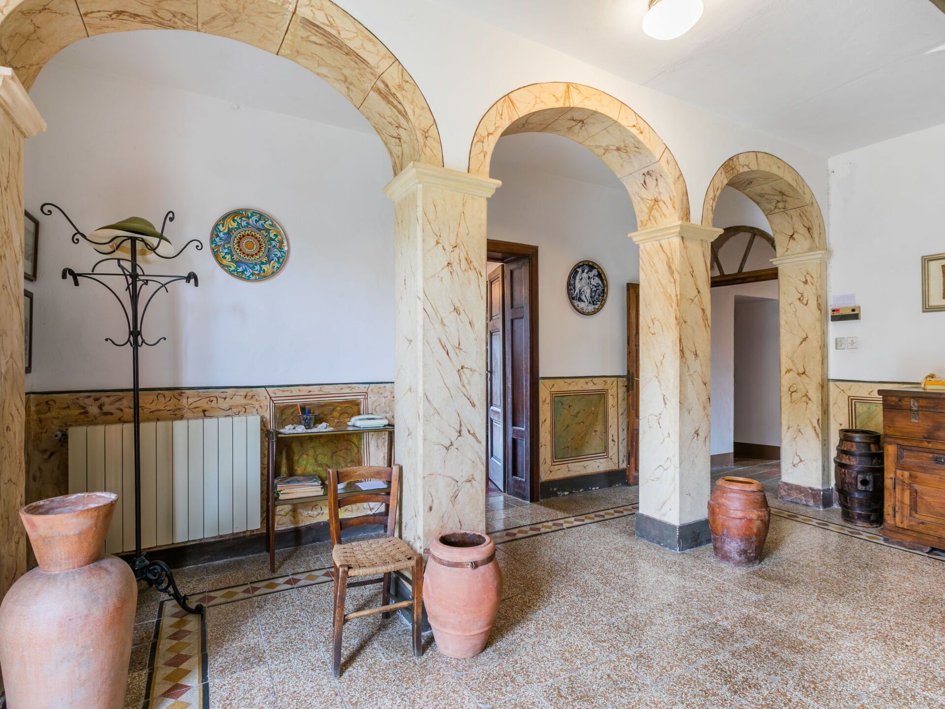Une pièce avec des arcades en marbre, un radiateur, une chaise, une table et une assiette décorative accrochée au mur.