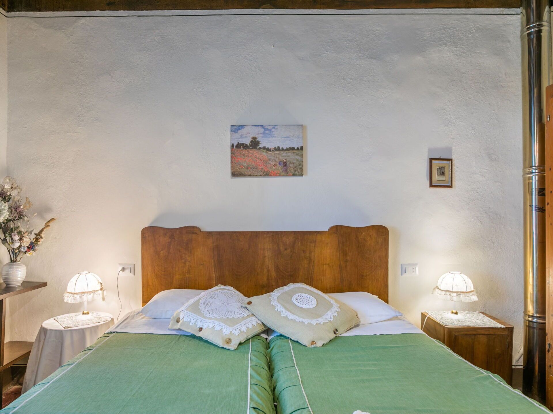 A bedroom with a wooden headboard, two bedside tables with lamps, a green bedspread, and a painting on the wall.