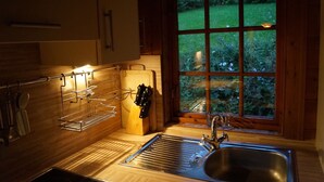 Una cocina con un lavamanos de acero inoxidable, armarios de madera, un bloque de cuchillos y una ventana con vistas a un jardín.