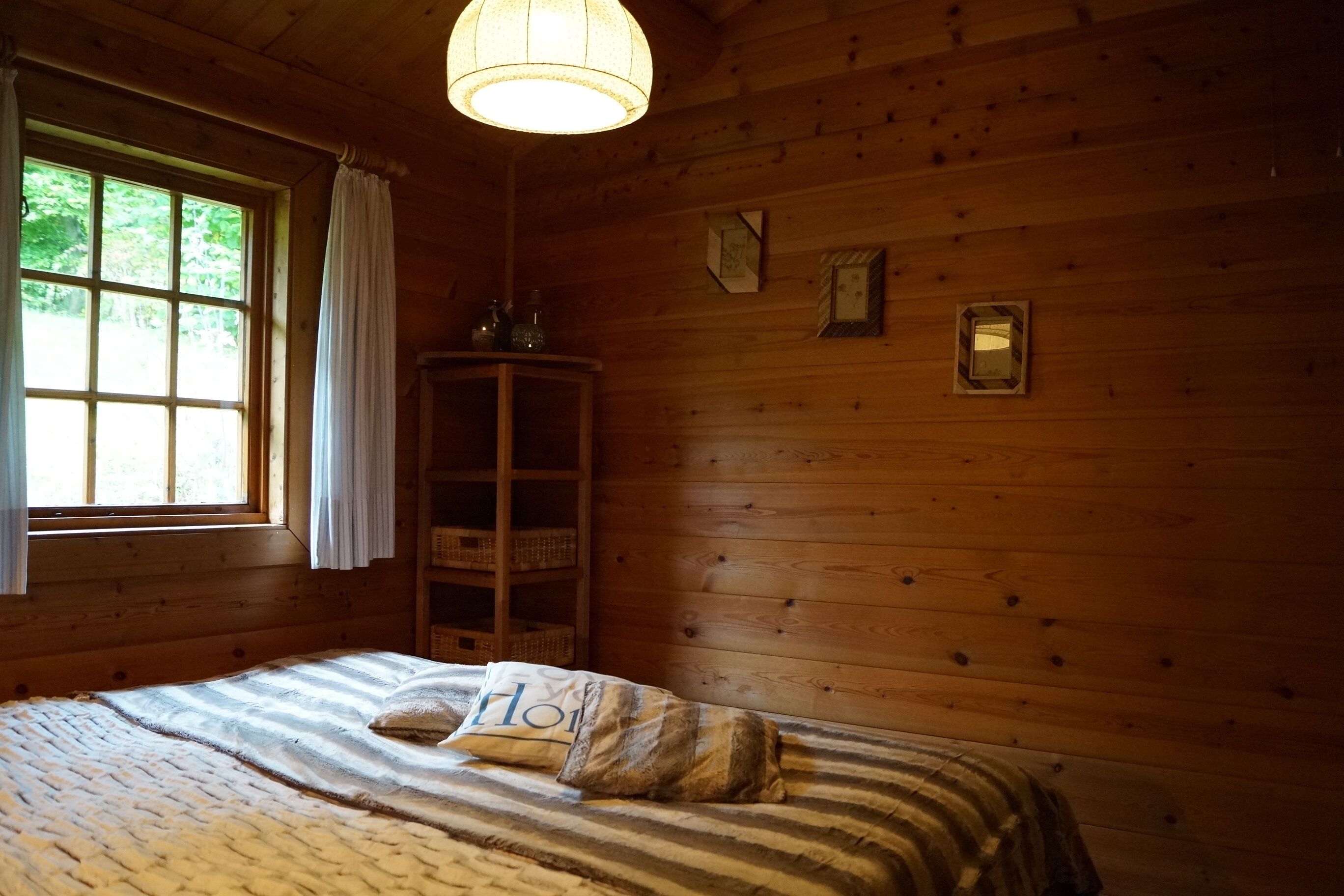 A wooden cabin room with a bed, a window with curtains, a wooden cabinet, and a hanging light fixture.